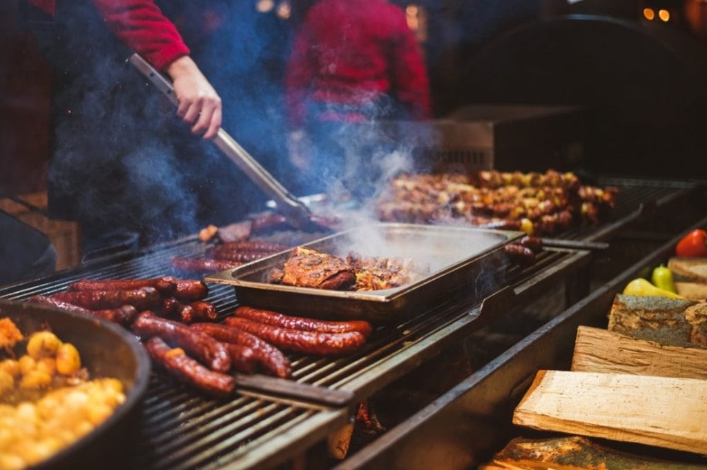 A person operating a BBQ. There is food cooking on the top of the grill