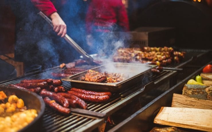 A person operating a BBQ. There is food cooking on the top of the grill