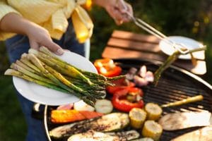 A woman is handing the camera man a plate of asparagus, while cooking other vegetables on a grill