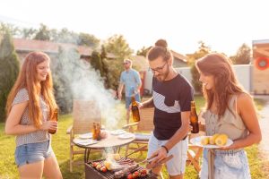 A group, one man and 2 women, are standing around the BBQ. There is beers and food, its a sunny day. 
