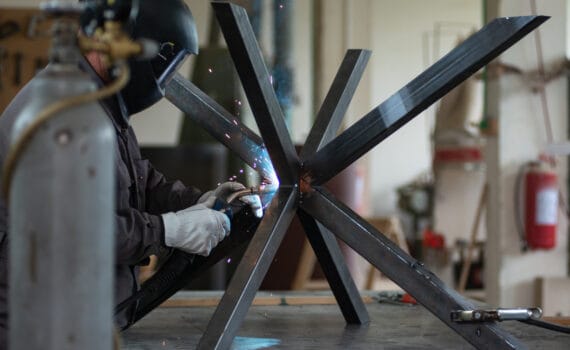 A mature blacksmith welds the metal structure of a dining table in a small workshop